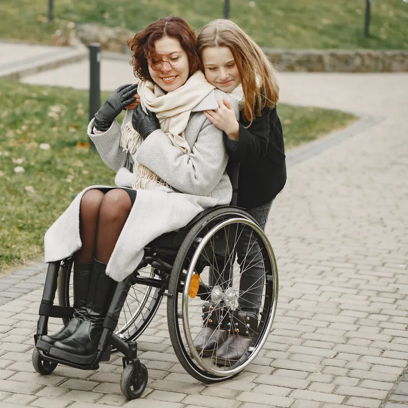 Mulher sorridente em cadeira de rodas manual abraçada pela filha em um parque